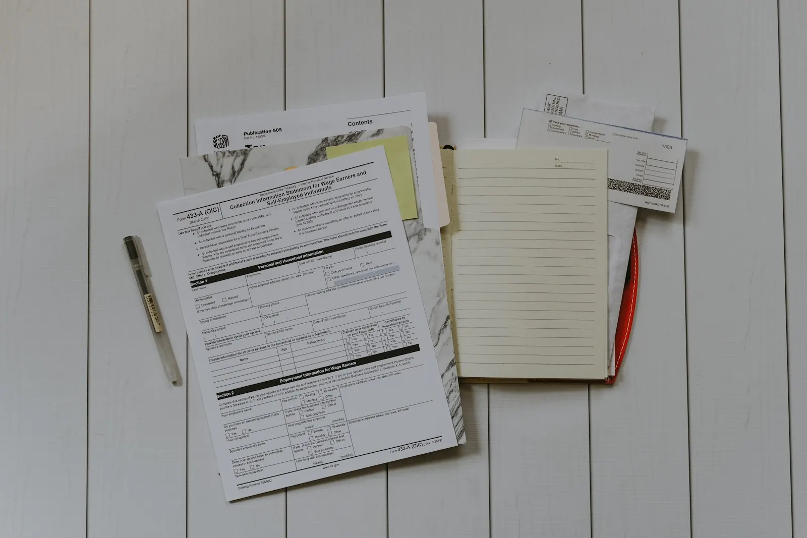 Flat lay of printed forms, a notepad and pens on a white wooden surface